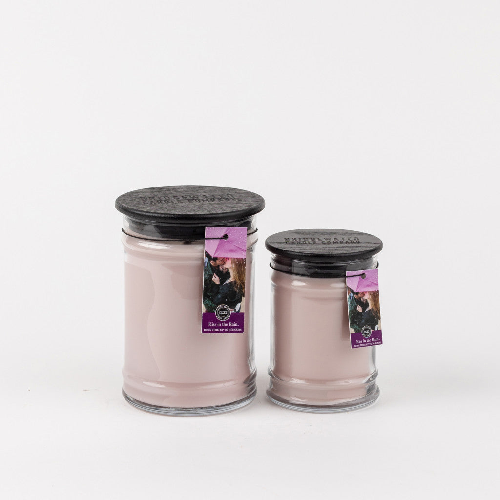 Two Kiss in the Rain 18oz Large Jar Candles by Bridgewater Candle Company, with light pink wax, black lids, and purple tags showing an image and text, are placed side by side on a plain white background.