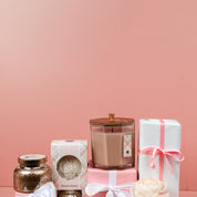 A gift set featuring a glass candle, Bridgewater Candle Companys Sweet Grace Mercury Flower Diffuser, a boxed flower, and two presents with pink and white ribbons, all beautifully arranged on a pink background.