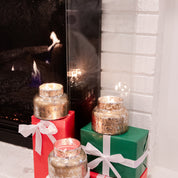 Three Sweet Grace Mercury Jar Candles by Bridgewater Candle Company, filled with soy-blend wax, are displayed on festive red and green gift boxes with white ribbons near a glowing fireplace and a white brick wall in the background.