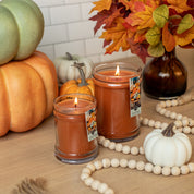 Two lit Bridgewater Candle Company Harvest Pumpkin 18oz Large Jar Candles sit on a wooden table with decorative pumpkins, wooden beads, and autumn leaves. White tile and seasonal accents appear in the background.