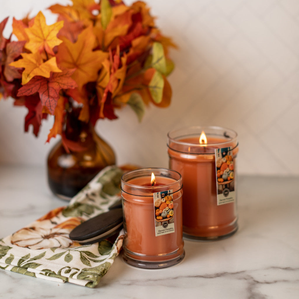 Two lit Bridgewater Candle Company Harvest Pumpkin 8oz Jar Candles rest on marble beside a brown vase of autumn leaves and a floral cloth, radiating a cozy fall ambiance with their inviting pumpkin scent.