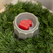 The Bridgewater Candle Company Wax Warmer-Grey Banded holds two red wax melts and sits amid a green pine wreath, creating a festive holiday display ideal for enjoying your favorite home fragrance oils.