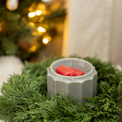 The Bridgewater Candle Company Christmas Bliss Flameless Bundle features red wax melts in a gray holder surrounded by pine branches, filling the air with festive fragrance as soft yellow holiday lights glow in the background.
