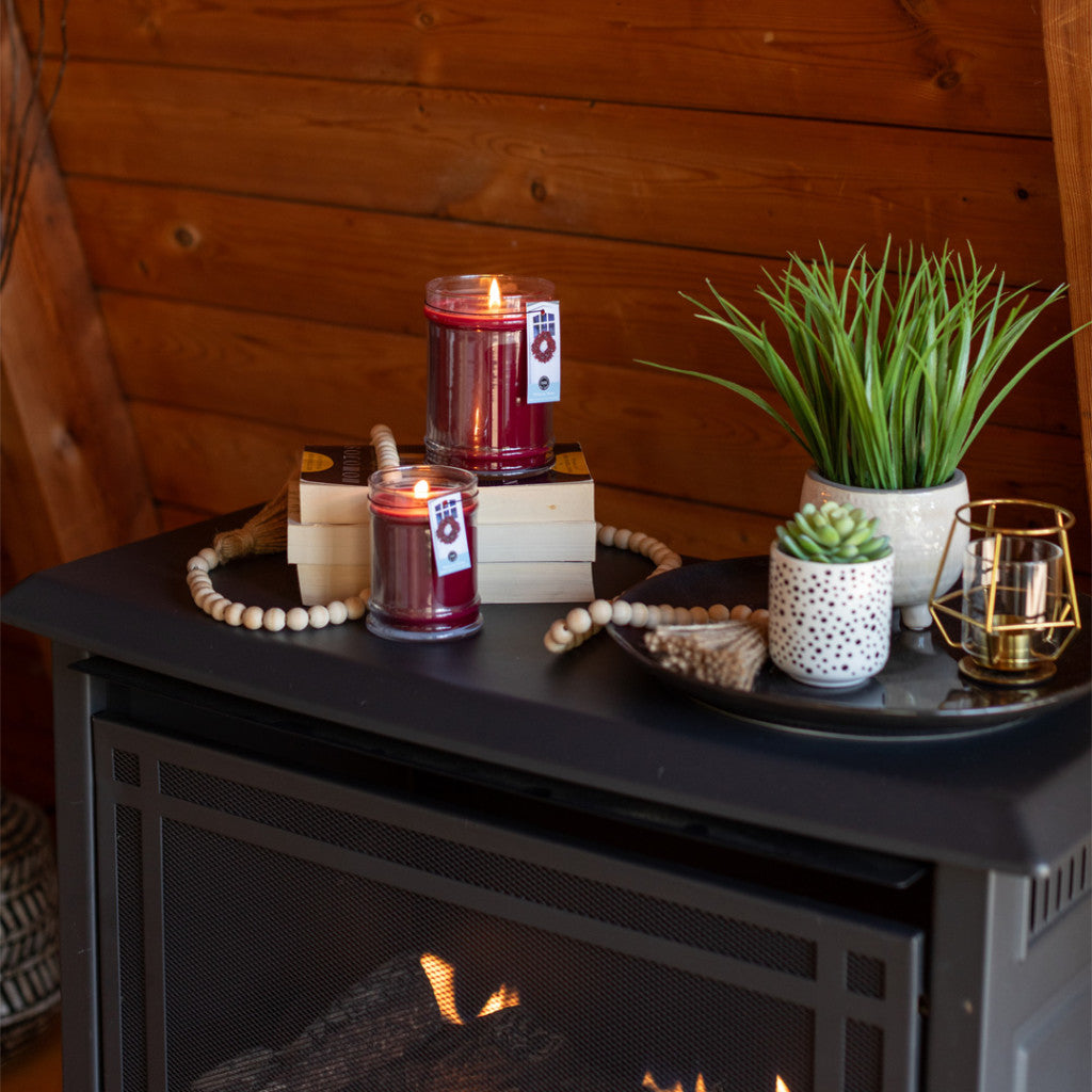 Two lit candles, including the Bridgewater Candle Company Welcome Home 18oz Large Jar Candle, sit on stacked books beside a potted plant and bead garland, all styled on a black mantel to create a cozy atmosphere by the fireplace.