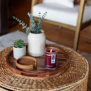 A woven round table displays a wooden tray with a small potted succulent, a larger vase with greenery, two woven coasters, and a lit Welcome Home 8oz Jar Candle by Bridgewater Candle Company. A chair with a white cushion is in the background.
