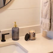 A bathroom countertop with a soap dispenser, a lit Bridgewater Candle Company Afternoon Retreat 8oz Jar Candle with its lid off, a black faucet, and two beige towels hanging on the wall. The edge of a round mirror is visible above.