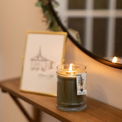 A Festive Frasier 8oz Jar Candle by Bridgewater Candle Company sits on a wooden shelf near a round mirror and framed gazebo sketch, its Pine Forest scent filling the cozy space with warm, inviting ambiance.