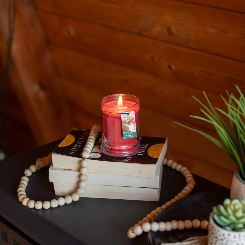 A Christmas Bliss 8oz Holiday Jar Candle by Bridgewater Candle Company burns atop two stacked books, including Four Thousand Weeks, next to a wooden bead garland and small green plant, set before a cozy wooden wall backdrop.