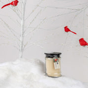 A Comfort & Joy 8oz Small Jar Candle from Bridgewater Candle Company sits on snowy ground before a white, leafless tree with three red cardinal ornaments, its minimalist setting evoking cozy seasonal fragrance and warmth.