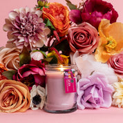 The XOXO 8oz Small Jar Candle by Bridgewater Candle Company, made with soy-blend wax, glows in a glass jar with a pink label, surrounded by artificial roses, dahlias, and peonies against a soft pink background.
