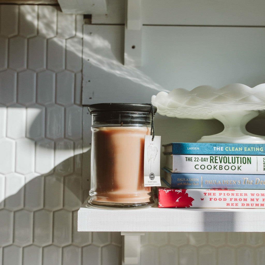 A Bridgewater Candle Company Sweet Grace 8oz Jar Candle sits on a white shelf near cookbooks and a cake stand, with sunlight casting shadows on a hexagonal tile backsplash.