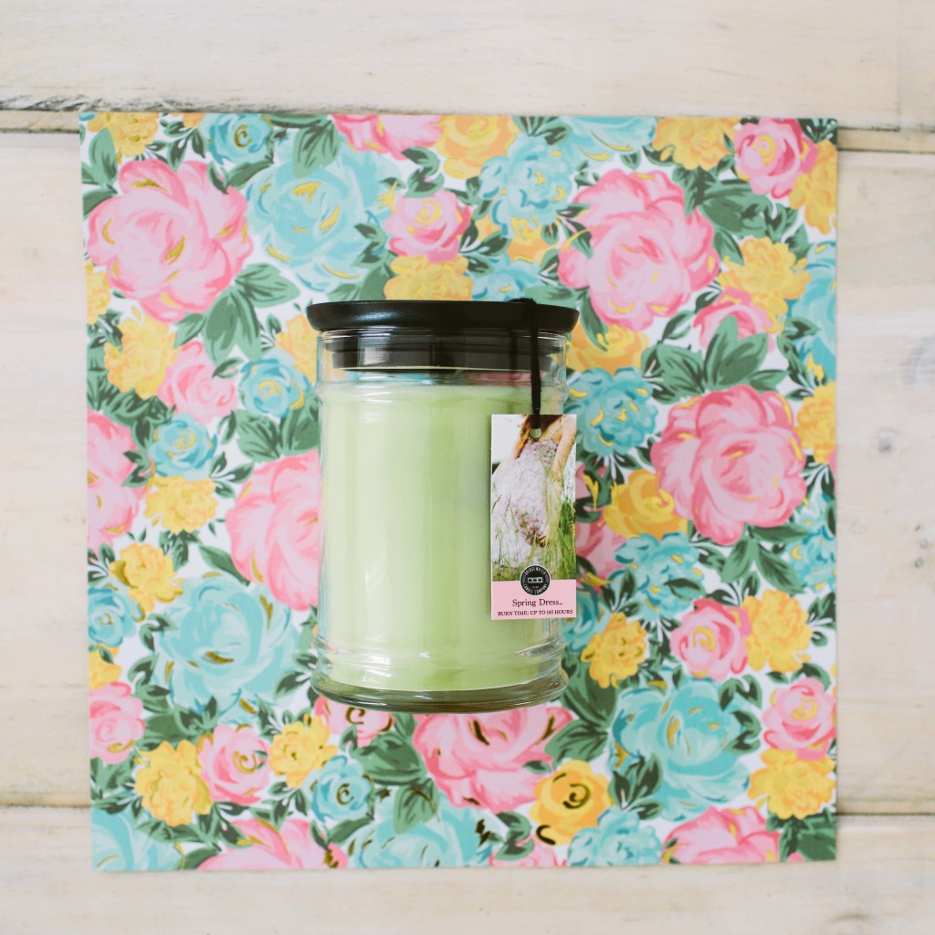 The Spring Dress 18oz Large Jar Candle by Bridgewater Candle Company, with a black lid, rests on a colorful floral square—featuring pink, yellow, and blue blooms—on light wood, perfect for adding fragrance to any room.