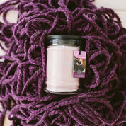 A Kiss in the Rain 18oz Large Jar Candle by Bridgewater Candle Company sits in a glass jar with black lid and tag on thick, chunky purple knit yarn.