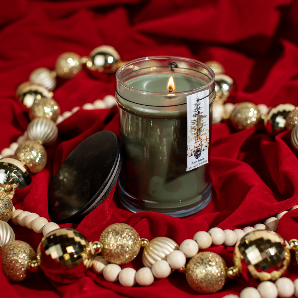 The Bridgewater Candle Company Festive Frasier 18oz Large Jar Candle sits on red fabric amid gold and white ornaments, its black lid nearby. Enjoy this signature scent for a cozy, festive atmosphere.