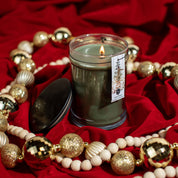 The Bridgewater Candle Company Festive Frasier 18oz Large Jar Candle sits on red fabric amid gold and white ornaments, its black lid nearby. Enjoy this signature scent for a cozy, festive atmosphere.