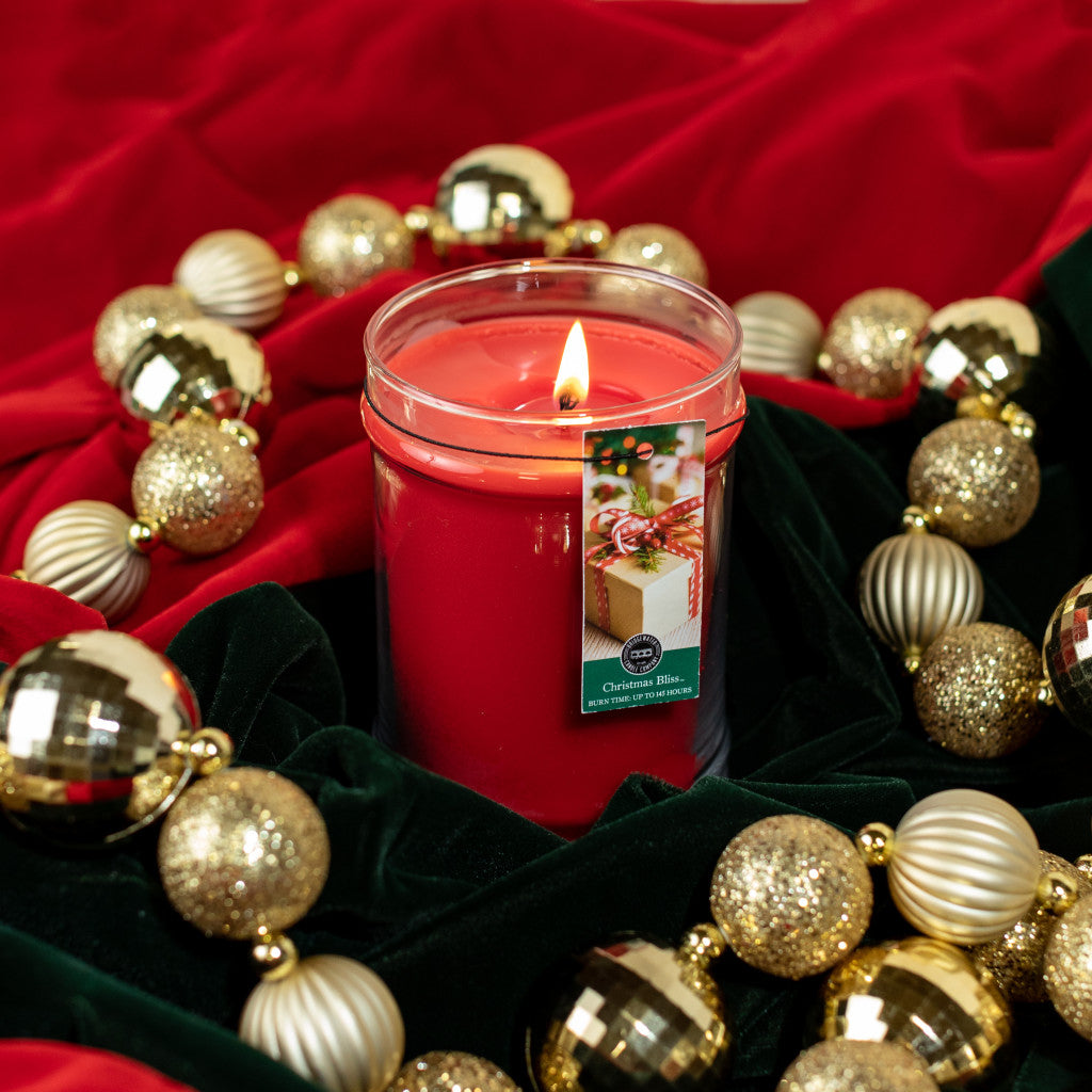 The Bridgewater Candle Company Christmas Bliss 18oz Large Jar burns brightly in glass, surrounded by gold and white ornaments on green and red velvet. Its label shows a festive present and holiday design.