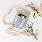 The Lavender Fields 18oz Large Jar Candle by Bridgewater Candle Company sits in a glass jar on a tray, accented by pink ribbon, gold scissors, lavender buds, and dried lavender against a white background.