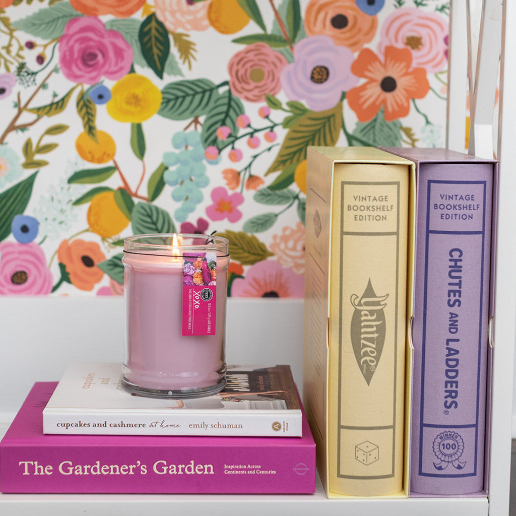 A lit XOXO 18oz Large Jar Candle by Bridgewater Candle Company sits in its glass jar on a stack of books beside three pastel hardcovers, set against vibrant floral wallpaper.