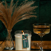 A lit Bridgewater Candle Company Amber & Emerald 18oz Large Jar Candle sits on a tray beside an ornate glass of amber liquid and a vase with dried pampas grass, set against a dark green background.