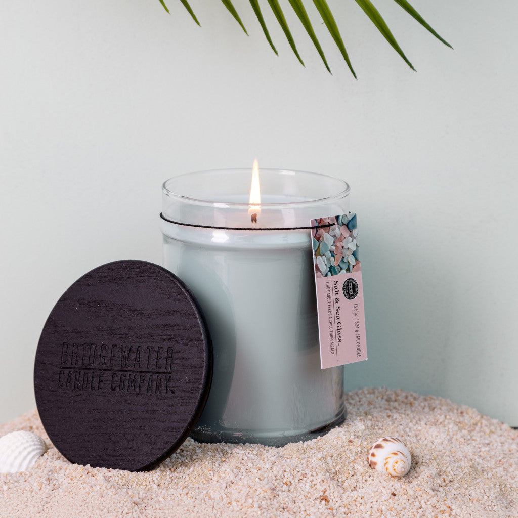The Bridgewater Candle Company Salt & Sea Glass 18 oz Large Jar Candle features a dark wooden lid, White Cotton tag, and soy-blend wax for a clean burn. Displayed on sand with seashells and palm leaves above, it evokes a tranquil, coastal vibe.