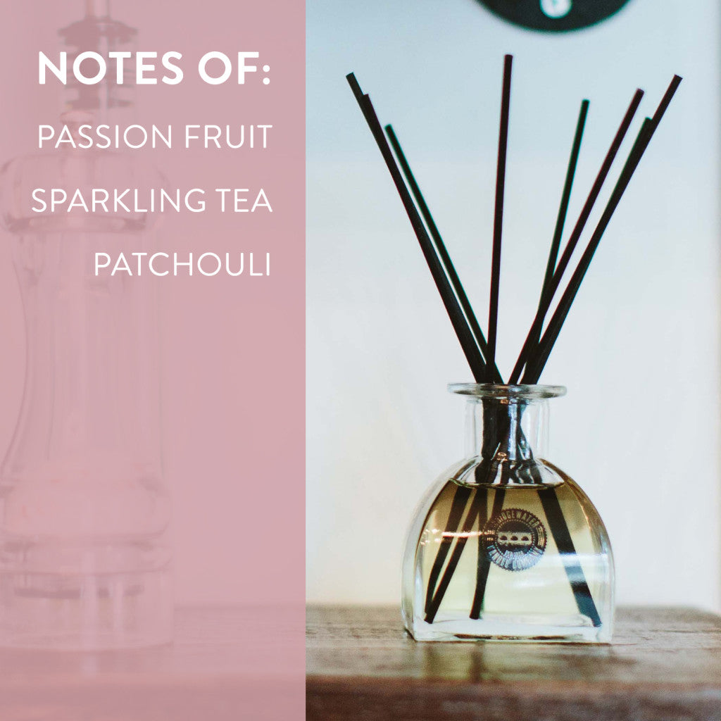 A Bridgewater Candle Company Sweet Grace Reed Diffuser sits on a wooden surface, filling the air with notes of passion fruit, sparkling tea, and patchouli. Black reeds add an elegant touch beside text on a pink background.