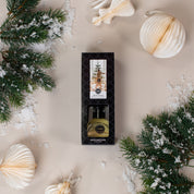 A boxed Festive Frasier Reed Diffuser by Bridgewater Candle Company, featuring a pine tree design, rests on a beige surface amid fiber reeds, faux evergreen branches, white paper ornaments, and scattered faux snow.
