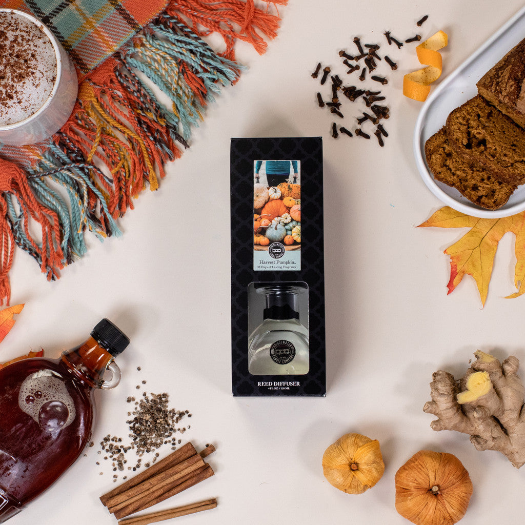 The Bridgewater Candle Companys Harvest Pumpkin Reed Diffuser, in a seasonal box, sits among pumpkin bread, spices, dried oranges, maple syrup, a plaid scarf, and autumn leaves—capturing the cozy essence of home fragrance for fall.