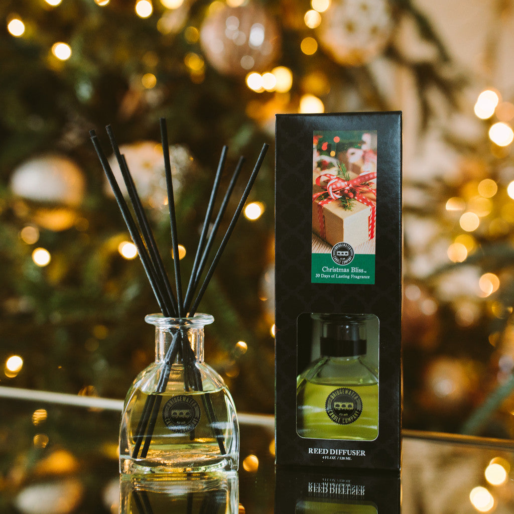 A Bridgewater Candle Company Christmas Bliss Reed Diffuser and box sit on a glass surface, backed by a softly blurred, glowing Christmas tree. Its light green fragrance brings festive charm to your holiday décor.