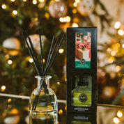 A Bridgewater Candle Company Christmas Bliss Reed Diffuser and box sit on a glass surface, backed by a softly blurred, glowing Christmas tree. Its light green fragrance brings festive charm to your holiday décor.