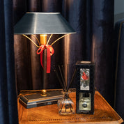 A black table lamp with a red ribbon sits on a wooden side table next to a Bridgewater Candle Company Comfort & Joy Reed Diffuser and boxed scented oil, creating an inviting home fragrance scene before dark blue curtains.