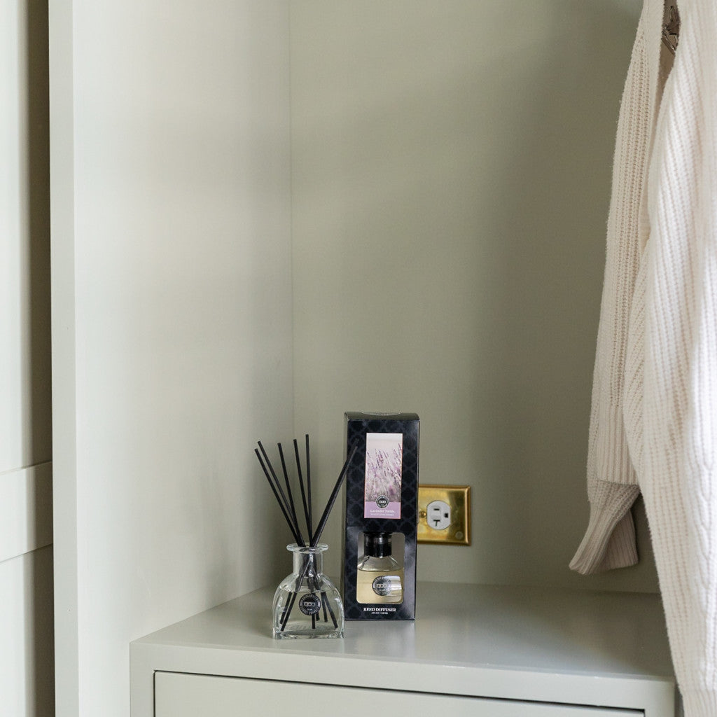 A Bridgewater Candle Company Lavender Fields Reed Diffuser sits on a light cabinet near an outlet, its fiber reeds dispersing fragrance. A boxed scent is nearby, and a cream textured garment hangs partially to the right.