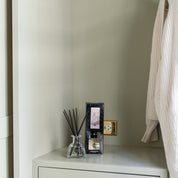 A Bridgewater Candle Company Lavender Fields Reed Diffuser sits on a light cabinet near an outlet, its fiber reeds dispersing fragrance. A boxed scent is nearby, and a cream textured garment hangs partially to the right.