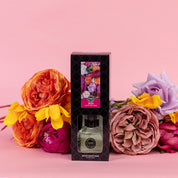 A Bridgewater Candle Company XOXO Reed Diffuser in a black, floral-designed box stands before vibrant artificial pink, purple, orange, and yellow flowers. Fiber reeds fill the air with fragrance against a pastel pink background.
