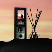 A Hello Night Reed Diffuser by Bridgewater Candle Company and its box are shown against a pink and yellow sunset. The box displays a person sitting on a car roof, gazing at the sky, as fiber reeds diffuse the premium fragrance.