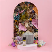 A gray ceramic diffuser and the Bridgewater Candle Company Sweet Grace Home Fragrance Oil sit on a white platform, in front of an arch with vibrant dried flowers, paper fans, and disco balls—perfect for flameless fragrance in your space.