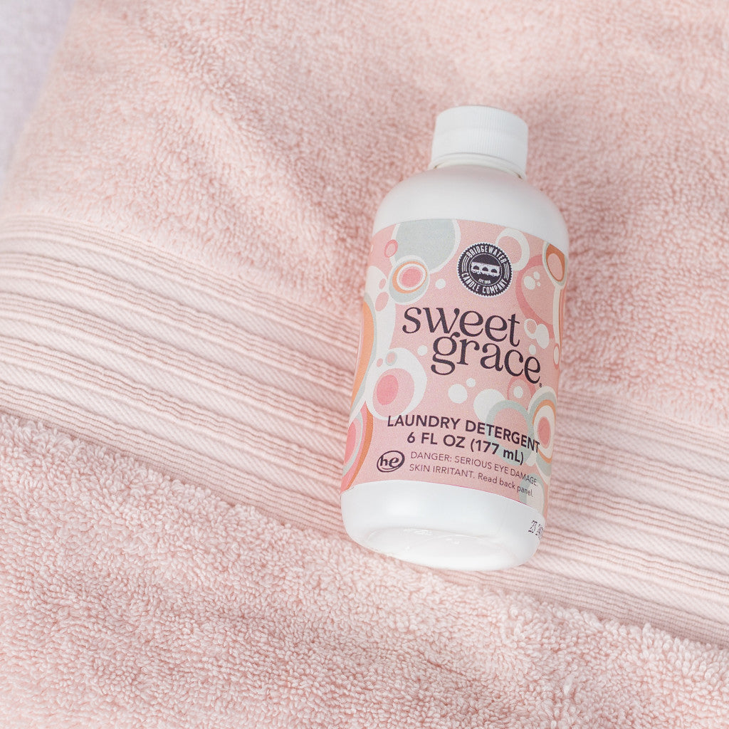 A bottle of Bridgewater Candle Company’s Sweet Grace 6oz Laundry Detergent sits on a soft pink towel, its pastel circular label highlighting a fabric-safe formula and long-lasting scent with clear black and white details.