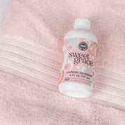 A bottle of Bridgewater Candle Company’s Sweet Grace 6oz Laundry Detergent sits on a soft pink towel, its pastel circular label highlighting a fabric-safe formula and long-lasting scent with clear black and white details.