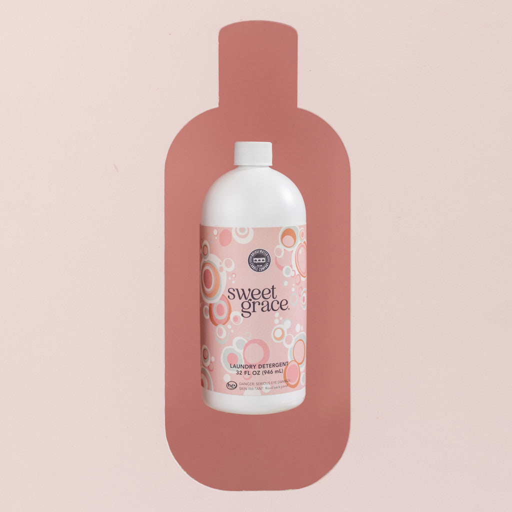 A white bottle of Bridgewater Candle Company’s Sweet Grace 32oz Laundry Detergent, with a pink bubbly label and dark pink silhouette, stands against a light pink background, promising a long-lasting scent.