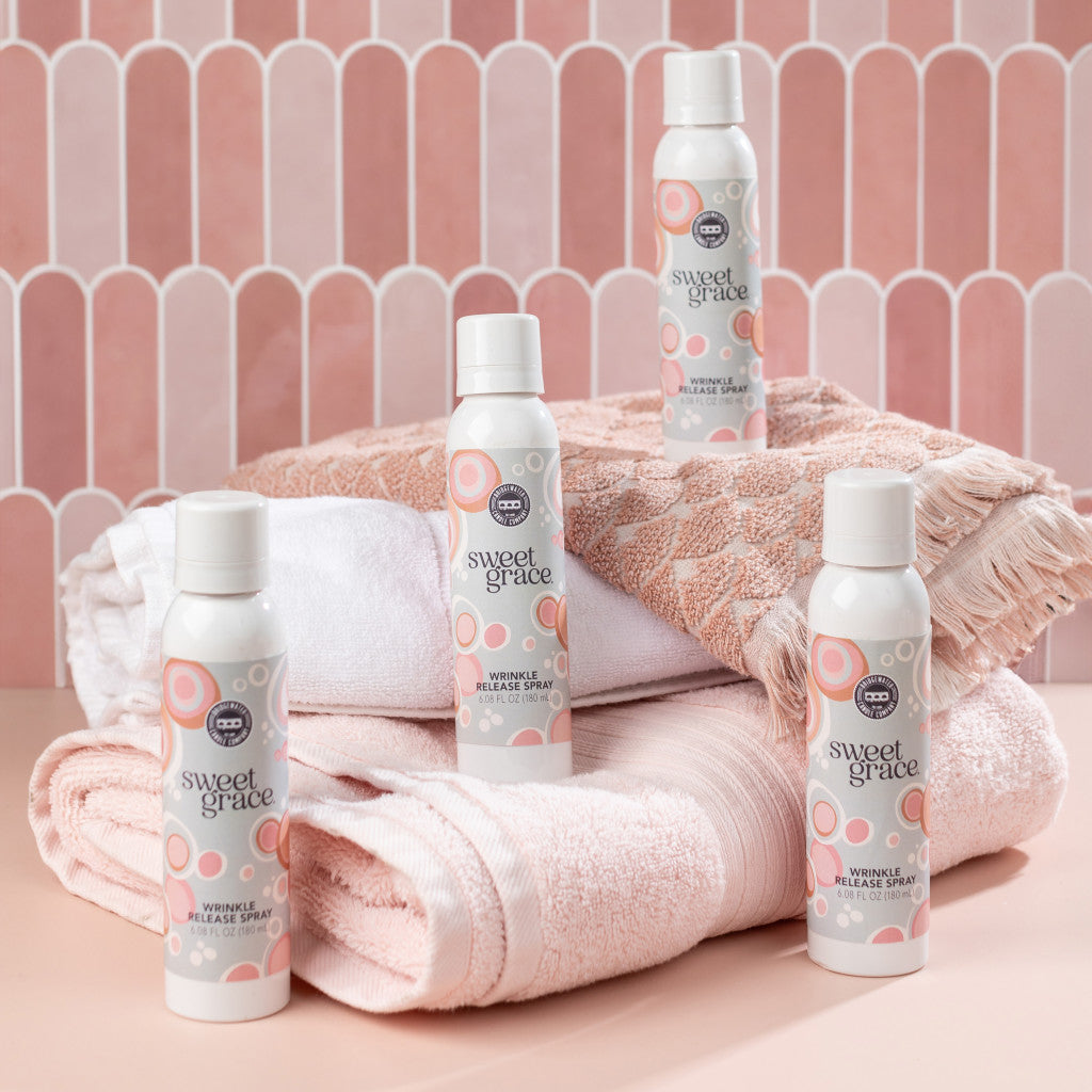 Four bottles of Bridgewater Candle Company’s Sweet Grace Wrinkle Release Spray sit on folded pink and white towels, set against a pink patterned background for a soft, clean, cozy laundry look.