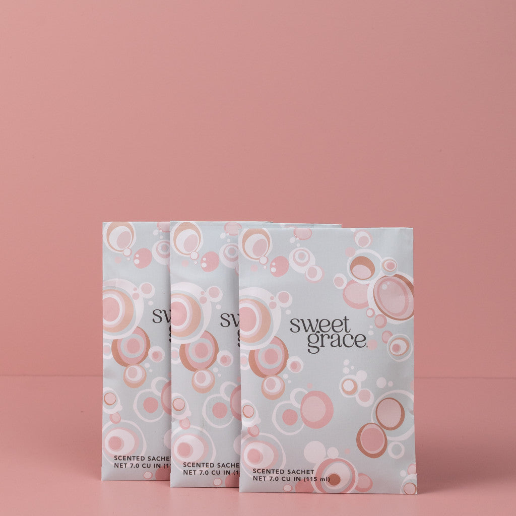 Three Bridgewater Candle Company Sweet Grace Modern Bubble Sachets, featuring a pink and white design, are lined up on a solid pink background—ideal for freshening up any room with delightful fragrance.