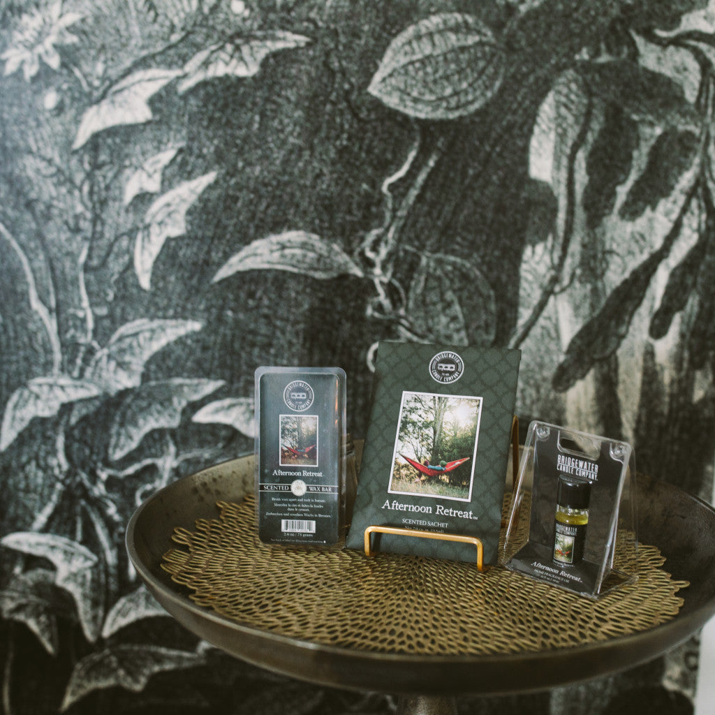 Three Bridgewater Candle Company Afternoon Retreat Flameless Bundle wax melts are displayed on a round table against leafy monochrome wallpaper, with the center package showing a red hammock in the woods.