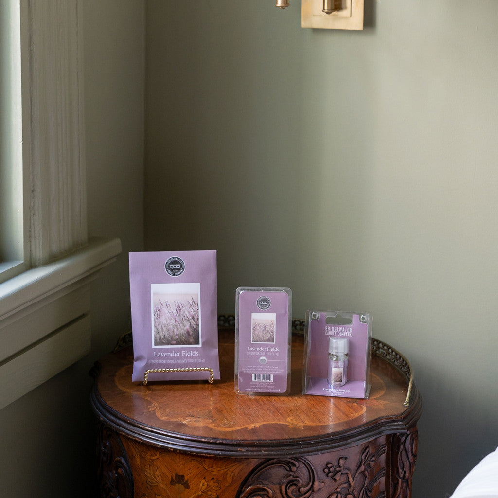 A wooden side table by the window displays the Bridgewater Candle Company’s Lavender Fields Flameless Bundle, featuring a booklet on a stand, a clamshell wax melt, and a small spray—all arranged neatly.