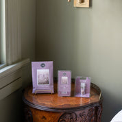 A wooden side table by the window displays the Bridgewater Candle Company’s Lavender Fields Flameless Bundle, featuring a booklet on a stand, a clamshell wax melt, and a small spray—all arranged neatly.