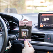 A hand holds a Bridgewater Candle Company Spring Dress Auto Vent Clip in front of a car dashboard displaying “Blank Space” by Taylor Swift, with sunlight streaming through the windshield.