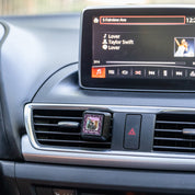 Car dashboard with a Bridgewater Candle Company Kiss in the Rain Auto Vent Clip and a display screen showing Lover by Taylor Swift playing, featuring her photo. The vent clip displays the Kiss in the Rain decorative image.