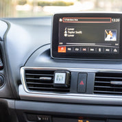 Car dashboard view with a media display playing Lover by Taylor Swift, album cover visible, navigation info at the top, and a Bridgewater Candle Company Laundry Line Auto Vent Clip adding freshness to the drive.