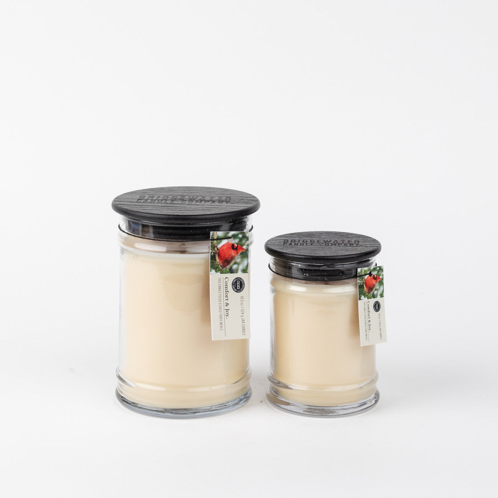 Two Bridgewater Candle Company glass jars, one being the Comfort & Joy 18oz Large Jar Candle, both with cream wax, black lids, and strawberry labels, are displayed against a white background.