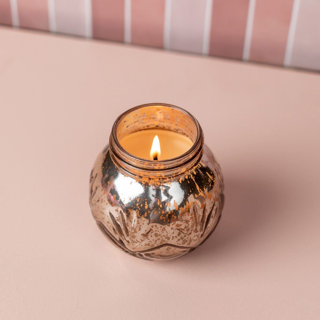 A lit Sweet Grace Collection Candle #008 by Bridgewater Candle Company, made with soy-blend wax, glows in a decorative metallic glass jar on a light pink surface against a pink tiled wall.