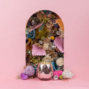 A metallic pink holder with the Bridgewater Candle Company’s Sweet Grace Collection Candle #011 sits before a floral arrangement of colorful dried flowers and disco balls, framed by an arched cutout against a pink backdrop.