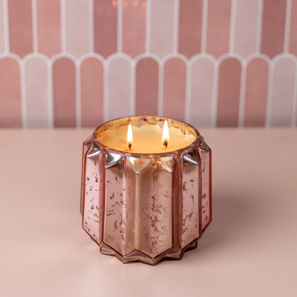 A lit Sweet Grace Collection Candle #021 by Bridgewater Candle Company with two flames glows in a faceted rose gold mercury glass holder of soy-blend wax, resting on a light pink surface against a pink and white tiled background.